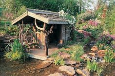 an outhouse in the middle of a stream surrounded by flowers and rocks, with a deer head sticking out of it's roof