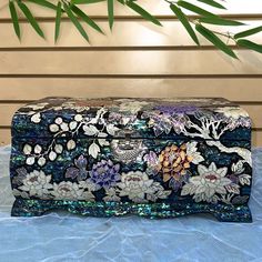 an ornately decorated box sitting on top of a blue cloth covered table next to a bamboo plant