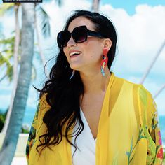 a woman wearing sunglasses and a yellow cardigan stands on the beach with palm trees in the background