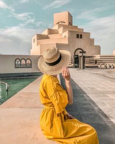 a woman in a yellow dress and hat sitting on the edge of a pool looking at an architectural building