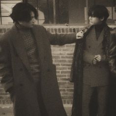 two men standing next to each other in front of a brick building and one man wearing a long coat