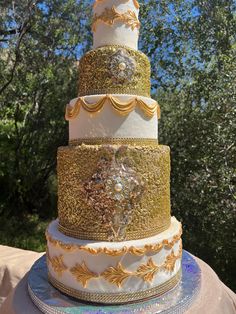 a three tiered wedding cake with gold and white decorations on the top, surrounded by trees