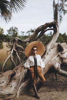 a man wearing a hat and sunglasses leaning against a tree trunk in front of the ocean