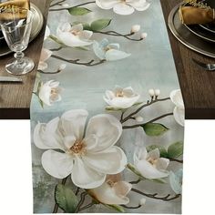 table runner with white flowers and green leaves on it, along with silver plates and utensils