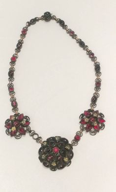 "This is a gorgeous older necklace made of antique brass with purple and red cabochon glass stones and little gold tone flowers. Most likely from the early 1900s to 1920s at the latest. It has been in my collection for many years. I can no longer wear it because it is too small for my neck. It has a locking clasp so in order for the necklace to be extended the clasp would have to be replaced and I just didn't want to do that. It is missing one red stone in front as pictured so I have priced it a Vintage Jeweled Necklace For Vintage Events, Antique Jeweled Red Jewelry, Vintage Ruby Necklace For Formal Occasions, Red Victorian Jewelry For Vintage Collection, Handmade Vintage Ruby Necklace, Red Ruby Victorian Necklace, Antique Jeweled Collectible Necklace, Antique Red Brass Necklace, Red Vintage Necklace For Collectors