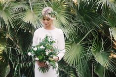 a woman standing in front of palm trees holding a bouquet of white and green flowers