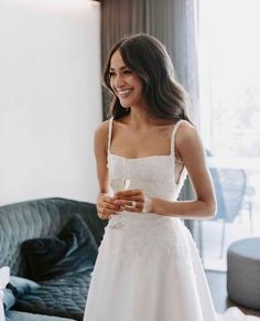 a woman in a white dress is smiling and looking at the camera while standing next to a couch