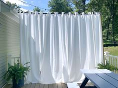 a white curtain hanging over a wooden deck next to a house with a bench and potted plant