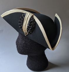 "This military style three cornered tricorn hat is made from top quality wool felt. The edges have been trimmed even and the sides held up or \"cocked\". It has been trimmed around the brim with gold fold-over braid. It is also available with black or white braid. Plain untrimmed hats are listed separately. Gold tone of edging may vary due to availability. Attached is a 3.5 \" black and gold, pleated double cockade with gold fleur de lis button below. Other buttons are available in the variation Pirate Hat With Curved Brim For Costume, Black Top Hat For Costume, Fitted Black Top Hat For Costume, Black Fitted Top Hat For Costume, Classic Black Hat For Costume, Adjustable Black Pirate Hat, Black Adjustable Pirate Hat, Formal Black Costume Hats With Curved Brim, Black Formal Costume Hat With Curved Brim