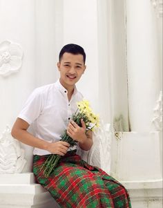 a man in a kilt sitting on a bench holding flowers and smiling at the camera