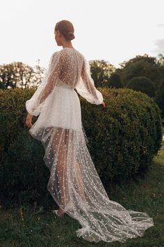 a woman in a white dress standing next to some bushes and grass with her back turned towards the camera