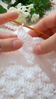 two hands are crocheting together on a white tablecloth with flowers in the background