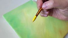 a person is holding a yellow paintbrush in their left hand and painting on a green piece of paper