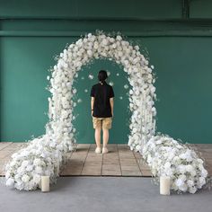 a person standing in front of a white flower arch with candles and flowers on the floor