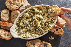 a person holding a piece of bread over a bowl of spinach artichoke dip