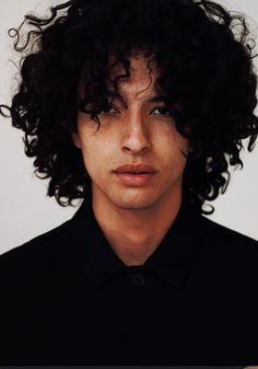 a man with curly hair and black shirt looking at the camera while wearing a watch