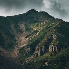 the top of a mountain with green grass on it's sides and dark clouds in the background