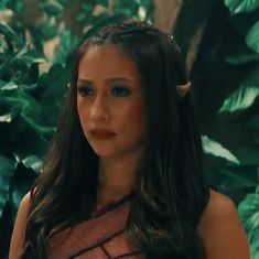 a woman with long hair and horns on her head standing in front of green plants