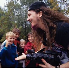 a man with long hair holding a camera in front of some young boys and girls