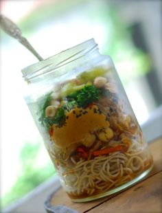 a jar filled with noodles and vegetables on top of a wooden table next to a window