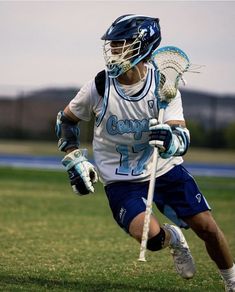 a lacrosse player running with the ball in his hand and wearing blue shorts, white shirt and black helmet