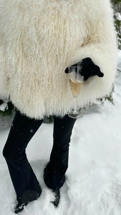 a person in black boots and a white sheep coat holding a glass of wine while standing in the snow