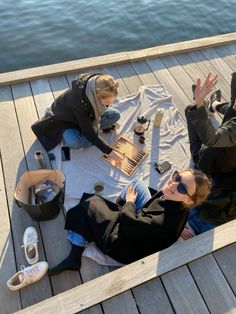 two people sitting on a dock with their hands in the air and one person holding out her hand