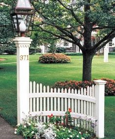 a white picket fence in front of a tree with flowers around it and a lamp post