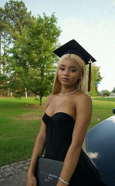 a woman in a graduation gown holding a book and standing next to a parked car