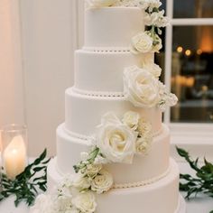 a wedding cake with white flowers and greenery on the table next to candlelight