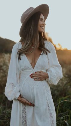 a pregnant woman in a white dress and cowboy hat smiles at the camera with her hands on her stomach