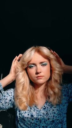 a woman with long blonde hair and blue eyeshades is holding her hands on her head