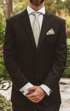 a man wearing a suit and tie standing in front of some bushes with his hands on his hips