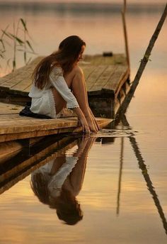 a woman is sitting on the edge of a dock looking at her reflection in the water