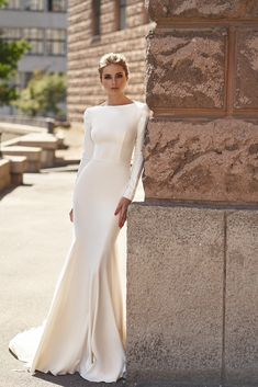 a woman in a long white dress leaning against a stone wall with her hands on her hips