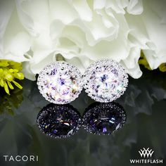 two diamond earrings sitting on top of a black surface next to flowers and white roses