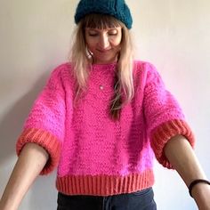 a woman wearing a pink sweater and green hat with her hands in the air while standing next to a white wall