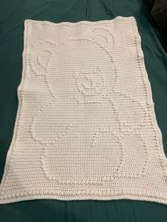 a white crocheted teddy bear blanket on a green bed sheet with a black pillow behind it