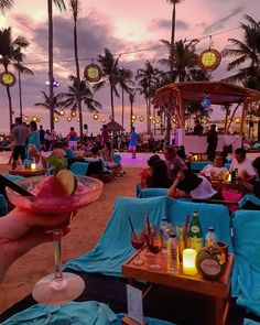 people sitting on the beach at sunset with drinks in their hands and lights hanging from palm trees