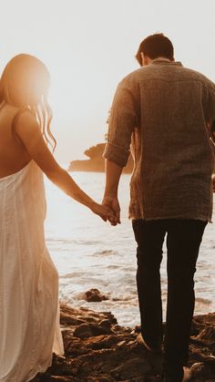 a man and woman holding hands while walking along the beach at sunset with the ocean in the background