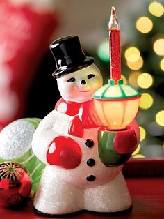 a snowman holding a lit candle next to a christmas ornament on a table