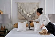 a woman in white shirt and black pants sitting on bed with pillows, hat and wall hanging