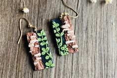 two earrings with green leaves on them sitting on a wooden table next to flowers and beads