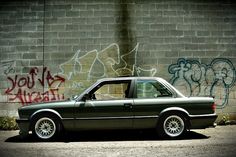 a black car parked in front of a brick wall with graffiti on it's side