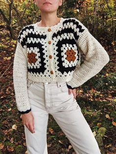 a woman is standing in the woods wearing a crocheted sweater and white jeans