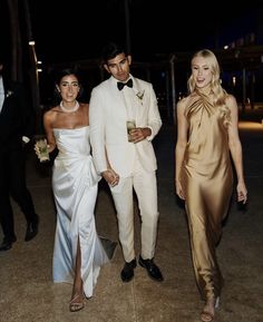 three people in formal wear standing next to each other on the sidewalk at night time