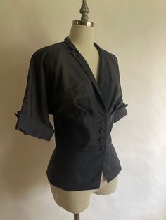 "1940s lightweight short sleeve jacket/blouse with pleated bust, covered buttons and lightly padded shoulders. Although the jacket appears to be black in the pictures, it is more of a slate grayMade by Wilshire of Boston, in excellent condition. The fabric is a bit of a mystery, it is lightweight with a striated texture, it could possibly be cotton or a form of rayon.  Shoulders 17\" across Chest 42\" (21\" across, doubled) Waist 34\" Hem 42\" Sleeve from neck to rolled cuff16\" Length from back of neck to hem 23\"" Formal Fitted Blouse With Lapel Collar, Fitted Formal Blouse With Buttons, Formal Fitted Blouse With Buttons, Semi-formal Fitted Tops With Covered Buttons, Fitted Short Sleeve Blouse For Semi-formal Events, Fitted Short Sleeve Blouse For Semi-formal Occasions, Fitted Blouse With Lapel Collar And Hidden Buttons, Fitted Tops With Lapel Collar For Formal Occasions, Formal Fitted Blouse With Button Closure