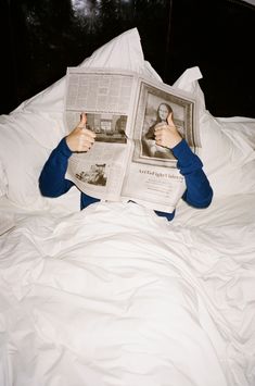 a person laying in bed reading a newspaper and pointing at the viewer with their thumb up