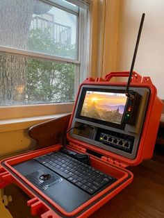 an orange laptop computer sitting on top of a wooden table next to a large window