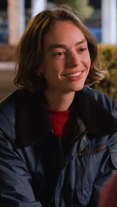 a smiling woman wearing a blue jacket and red shirt sitting in front of a building